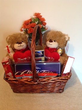 A wicker basket filled with an assortment of gifts including two plush teddy bears holding red heart-shaped pillows with 'I Love You' written on them. The basket also contains various chocolates such as Maltesers, Cadbury Milk Tray, and Kinder chocolates. A bouquet of orange roses sits behind the teddy bears.