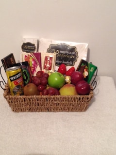 A wicker basket contains an assortment of items including chocolate bars, a can of energy drink, and a variety of fruits such as strawberries, grapes, green apples, and pears. The items are arranged neatly with chocolate packets and boxes standing upright at the back.