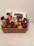 A wicker basket contains an assortment of items including chocolate bars, a can of energy drink, and a variety of fruits such as strawberries, grapes, green apples, and pears. The items are arranged neatly with chocolate packets and boxes standing upright at the back.