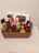 A wicker basket contains an assortment of items including chocolate bars, a can of energy drink, and a variety of fruits such as strawberries, grapes, green apples, and pears. The items are arranged neatly with chocolate packets and boxes standing upright at the back.