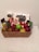 A wicker basket contains an assortment of items including chocolate bars, a can of energy drink, and a variety of fruits such as strawberries, grapes, green apples, and pears. The items are arranged neatly with chocolate packets and boxes standing upright at the back.