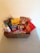 A wicker basket filled with various packaged candies and chocolates, including brands like Starburst, Kinder, and Allen's. The basket is placed on a light-colored surface with a plain background.