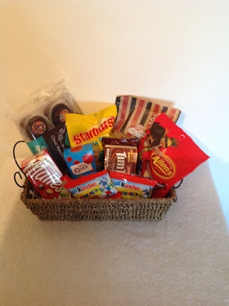 A wicker basket filled with various packaged candies and chocolates, including brands like Starburst, Kinder, and Allen's. The basket is placed on a light-colored surface with a plain background.
