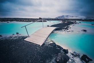 A small wooden bridge leads over a stream of striking turquoise water, surrounded by black volcanic rocks. The sky is overcast, creating a moody and serene atmosphere. In the distance, misty mountains and steam suggest geothermal activity.