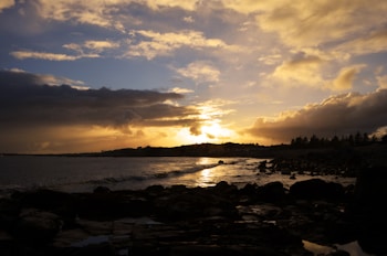 A coastal landscape at sunset with a dramatic sky. The sun is setting on the horizon, casting golden and orange hues across the sky and reflecting on the water. Dark clouds contrast with the warm colors of the sunset, creating a striking scene. In the foreground, rocky shores and waves are visible, while silhouettes of distant hills and trees line the horizon.