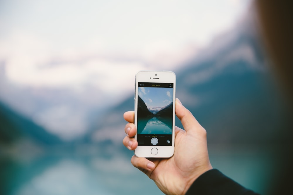 Person, die silbernes iPhone 5s locht und tagsüber ein Foto von einem Gewässer in der Nähe von Bergen macht