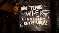 A black signboard with white painted text in Portuguese reads 'NÃO TEMOS WI-FI CONVERSEM ENTRE VOCÊS,' which translates to 'We don't have Wi-Fi, talk among yourselves.' The signboard is slightly worn, with a rustic appearance. There are decorative elements around it, along with some ambient lighting illuminating parts of the sign.