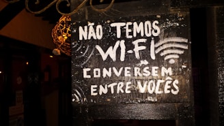A black signboard with white painted text in Portuguese reads 'NÃO TEMOS WI-FI CONVERSEM ENTRE VOCÊS,' which translates to 'We don't have Wi-Fi, talk among yourselves.' The signboard is slightly worn, with a rustic appearance. There are decorative elements around it, along with some ambient lighting illuminating parts of the sign.