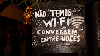 A black signboard with white painted text in Portuguese reads 'NÃO TEMOS WI-FI CONVERSEM ENTRE VOCÊS,' which translates to 'We don't have Wi-Fi, talk among yourselves.' The signboard is slightly worn, with a rustic appearance. There are decorative elements around it, along with some ambient lighting illuminating parts of the sign.