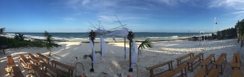 A serene beach setting featuring an altar decorated with flowers and white fabric, with wooden benches arranged on either side. The ocean waves gently roll onto the shore under a clear blue sky, while palm trees sway in the background, creating an inviting and tranquil ambiance.
