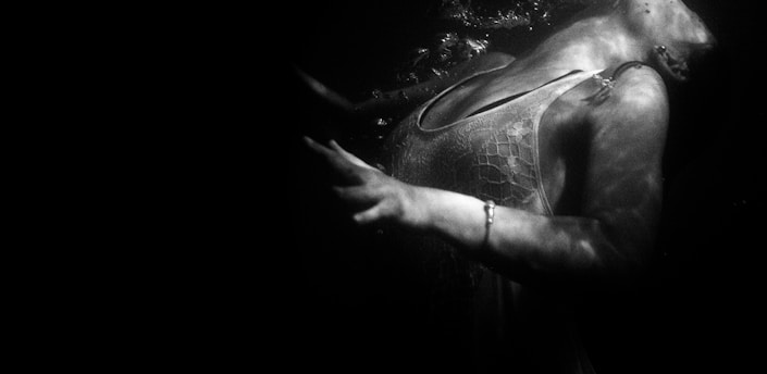 A black and white image of a person submerged underwater, partially revealing their upper body and left arm. Their face is turned away, with bubbles indicating movement. The lighting creates intricate shadows on the skin and clothing.