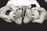 A close-up black and white photograph of baby feet being gently held by adult hands. The small toes are prominently displayed, capturing a moment of tenderness and care.
