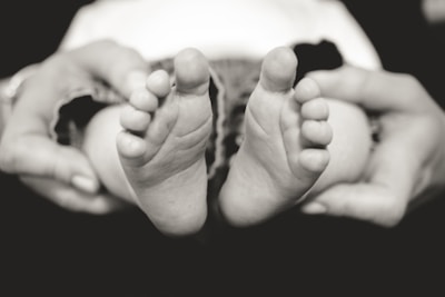 A close-up black and white photograph of baby feet being gently held by adult hands. The small toes are prominently displayed, capturing a moment of tenderness and care.