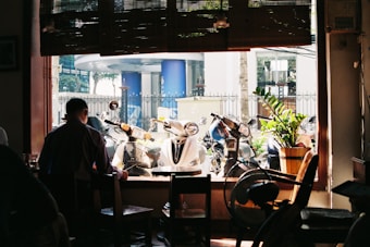 A cozy indoor cafe scene with a large window at the center, showing several parked scooters outside. A person is sitting at a table near the window, partially silhouetted against the daylight. A potted plant and a fan are visible inside, adding a touch of greenery and ventilation.