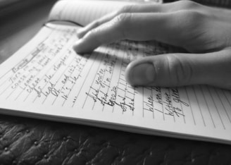 A close-up of a hand writing in a lined notebook, capturing a moment of handwriting with detailed focus on the fingers and penmanship. The image is in black and white, emphasizing the texture of the paper and the writing instrument.