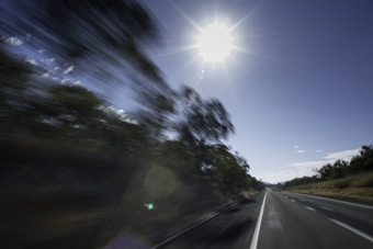 A dynamic scene featuring a highway with blurred trees on the side, indicating high speed. The sun shines brightly in the clear sky, casting harsh light and creating lens flare effects.
