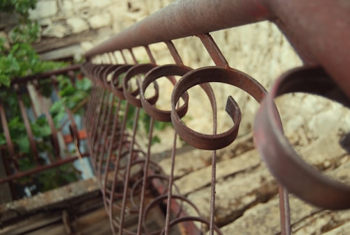 A rustic metal railing features a series of circular designs, extending into the distance with a slightly blurred background. The railing is weathered, with hints of rust, and it contrasts against the surrounding greenery and stone textures.