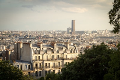 A cityscape featuring a classic European architecture with white buildings and mansard roofs in the foreground. A modern skyscraper is visible in the background under a partly cloudy sky, with lush green trees framing the scene.