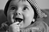 A close-up black and white photograph of a baby wearing a striped hat. The baby appears to be gazing upwards with an open-mouthed smile and has one hand near the mouth. The baby’s eyes are wide and expressive, highlighting the innocence and joy in its expression.