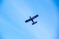 A small aircraft with a blue body and red accents flies against a clear blue sky. The aircraft is viewed from below, with its wings and tail clearly visible. The registration number can be seen on the side of the fuselage.