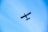 A small aircraft with a blue body and red accents flies against a clear blue sky. The aircraft is viewed from below, with its wings and tail clearly visible. The registration number can be seen on the side of the fuselage.