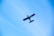 A small aircraft with a blue body and red accents flies against a clear blue sky. The aircraft is viewed from below, with its wings and tail clearly visible. The registration number can be seen on the side of the fuselage.