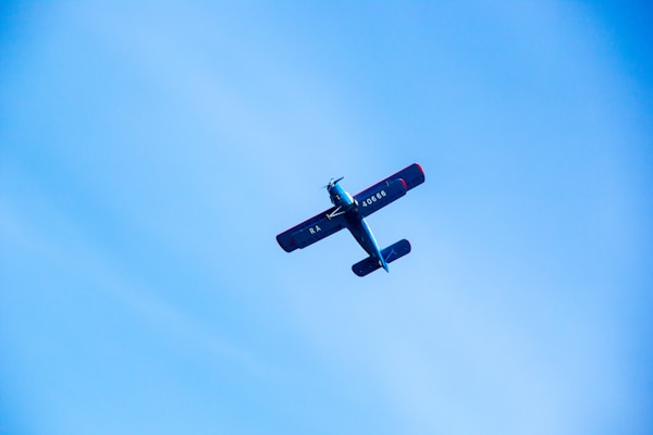 A small aircraft with a blue body and red accents flies against a clear blue sky. The aircraft is viewed from below, with its wings and tail clearly visible. The registration number can be seen on the side of the fuselage.