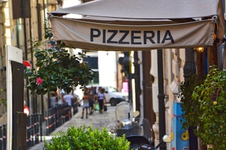 A narrow cobblestone street lined with old buildings features a pizzeria sign hanging above. Potted plants with vibrant flowers decorate the area, and a few pedestrians stroll in the background. A scooter is parked nearby, adding to the urban atmosphere.