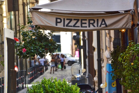 A narrow cobblestone street lined with old buildings features a pizzeria sign hanging above. Potted plants with vibrant flowers decorate the area, and a few pedestrians stroll in the background. A scooter is parked nearby, adding to the urban atmosphere.