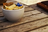 A small white bowl filled with individual packets of sugar sits on a wooden table. The packets are mostly yellow, with one blue packet visible. The table has a rustic texture with visible wood grain.