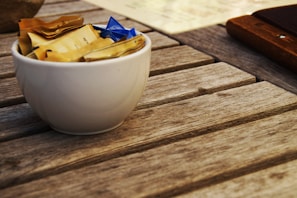 A small white bowl filled with individual packets of sugar sits on a wooden table. The packets are mostly yellow, with one blue packet visible. The table has a rustic texture with visible wood grain.