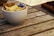 A small white bowl filled with individual packets of sugar sits on a wooden table. The packets are mostly yellow, with one blue packet visible. The table has a rustic texture with visible wood grain.