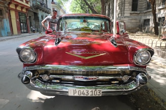 A vintage red Chevrolet car is parked on a street with a person standing next to it. The vehicle features a glossy finish, prominent headlights, and a classic Chevrolet emblem on the front. The background includes old buildings and trees lining the road, casting dappled shadows.