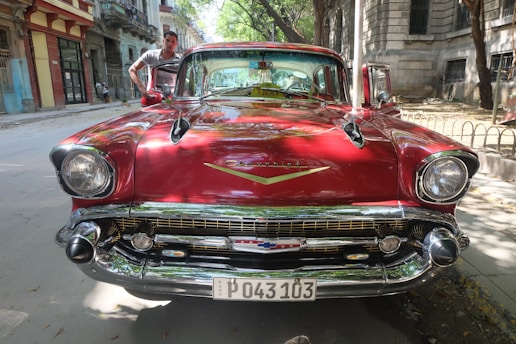 A vintage red Chevrolet car is parked on a street with a person standing next to it. The vehicle features a glossy finish, prominent headlights, and a classic Chevrolet emblem on the front. The background includes old buildings and trees lining the road, casting dappled shadows.