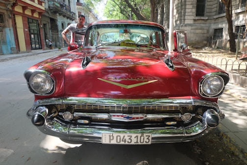 A vintage red Chevrolet car is parked on a street with a person standing next to it. The vehicle features a glossy finish, prominent headlights, and a classic Chevrolet emblem on the front. The background includes old buildings and trees lining the road, casting dappled shadows.