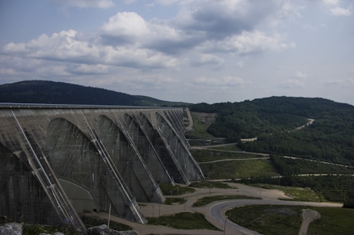 A large dam structure stretches across a landscape with rolling hills covered in greenery. The dam has a series of arched supports and is made of concrete. A winding road is visible in the foreground, leading towards the dam. The sky is overcast with scattered clouds, creating a dramatic backdrop.