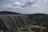 A large dam structure stretches across a landscape with rolling hills covered in greenery. The dam has a series of arched supports and is made of concrete. A winding road is visible in the foreground, leading towards the dam. The sky is overcast with scattered clouds, creating a dramatic backdrop.