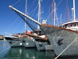 Several yachts are docked side by side in a marina, with their bows pointed slightly towards the viewer. They have wooden decks and netting at the front. The reflection of the water creates a shimmering effect on the sides of the boats. Masts and rigging rise into a clear blue sky.