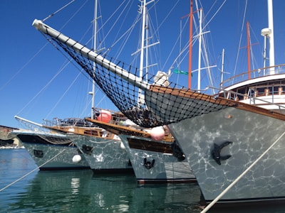 Several yachts are docked side by side in a marina, with their bows pointed slightly towards the viewer. They have wooden decks and netting at the front. The reflection of the water creates a shimmering effect on the sides of the boats. Masts and rigging rise into a clear blue sky.