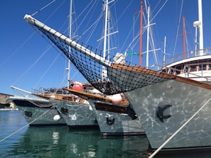 Several yachts are docked side by side in a marina, with their bows pointed slightly towards the viewer. They have wooden decks and netting at the front. The reflection of the water creates a shimmering effect on the sides of the boats. Masts and rigging rise into a clear blue sky.