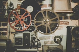 A vintage film projector is surrounded by large film reels and old records on a shelf. The color scheme includes muted tones, and the setting evokes a nostalgic, retro atmosphere. The background features a brick wall with some decorative items, such as a small tree and various gadgets that add to the antique theme.