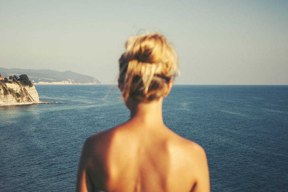 topless woman standing facing blue sea under white sky