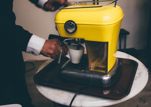 A person in a suit operates a vibrant yellow espresso machine, holding a small white cup under the coffee spout. The machine is placed on a round marble table and has a chrome base, giving it a retro style. The scene suggests a preparation of coffee.