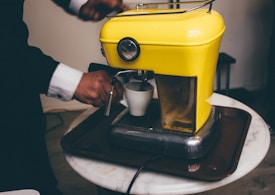 A person in a suit operates a vibrant yellow espresso machine, holding a small white cup under the coffee spout. The machine is placed on a round marble table and has a chrome base, giving it a retro style. The scene suggests a preparation of coffee.