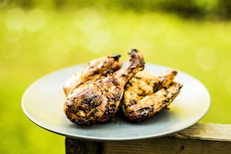 Three pieces of grilled chicken are placed on a round, light-colored plate. The chicken has a charred and seasoned appearance, suggesting it has been barbecued or grilled to a crispy texture. The background is blurred with a soft, green bokeh effect, which might indicate an outdoor setting.