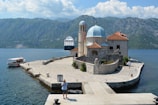 A picturesque stone island with a small historic church featuring a blue dome and terracotta roof is situated on calm blue waters. The island is surrounded by mountainous terrain under a partly cloudy sky. A cruise ship is seen in the distance, along with a tourist boat near the pier. People are exploring the island, creating a serene and scenic atmosphere.