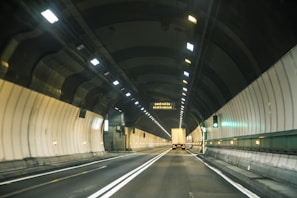 A brightly lit road tunnel with smooth walls and overhead ventilation ducts. Two vehicles, a truck and a car, are visible traveling through, with signage displaying information.