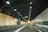 A brightly lit road tunnel with smooth walls and overhead ventilation ducts. Two vehicles, a truck and a car, are visible traveling through, with signage displaying information.