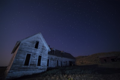 An old, weathered wooden house with peeling paint sits isolated under a clear, starry night sky. The structure appears abandoned, surrounded by barren land with distant hills. The scene is serene yet eerie.