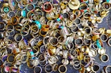 A large collection of various rings scattered on a dark surface. The rings display a variety of designs, with some featuring turquoise stones, intricate patterns, and metallic finishes. Their colors range from gold and silver to bright blues and reds, creating an eclectic and vibrant assortment.